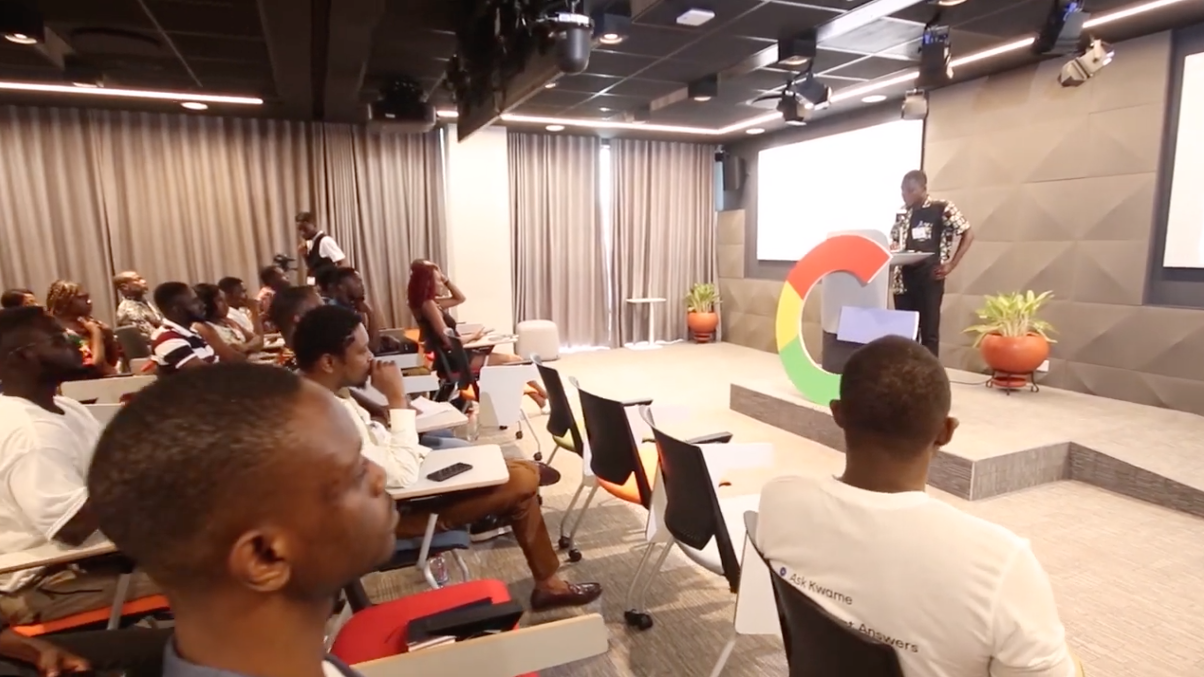 A grey high-tech looking conference room at the AfricAIED. A speaker stands on the right side in front of a seated audience. 