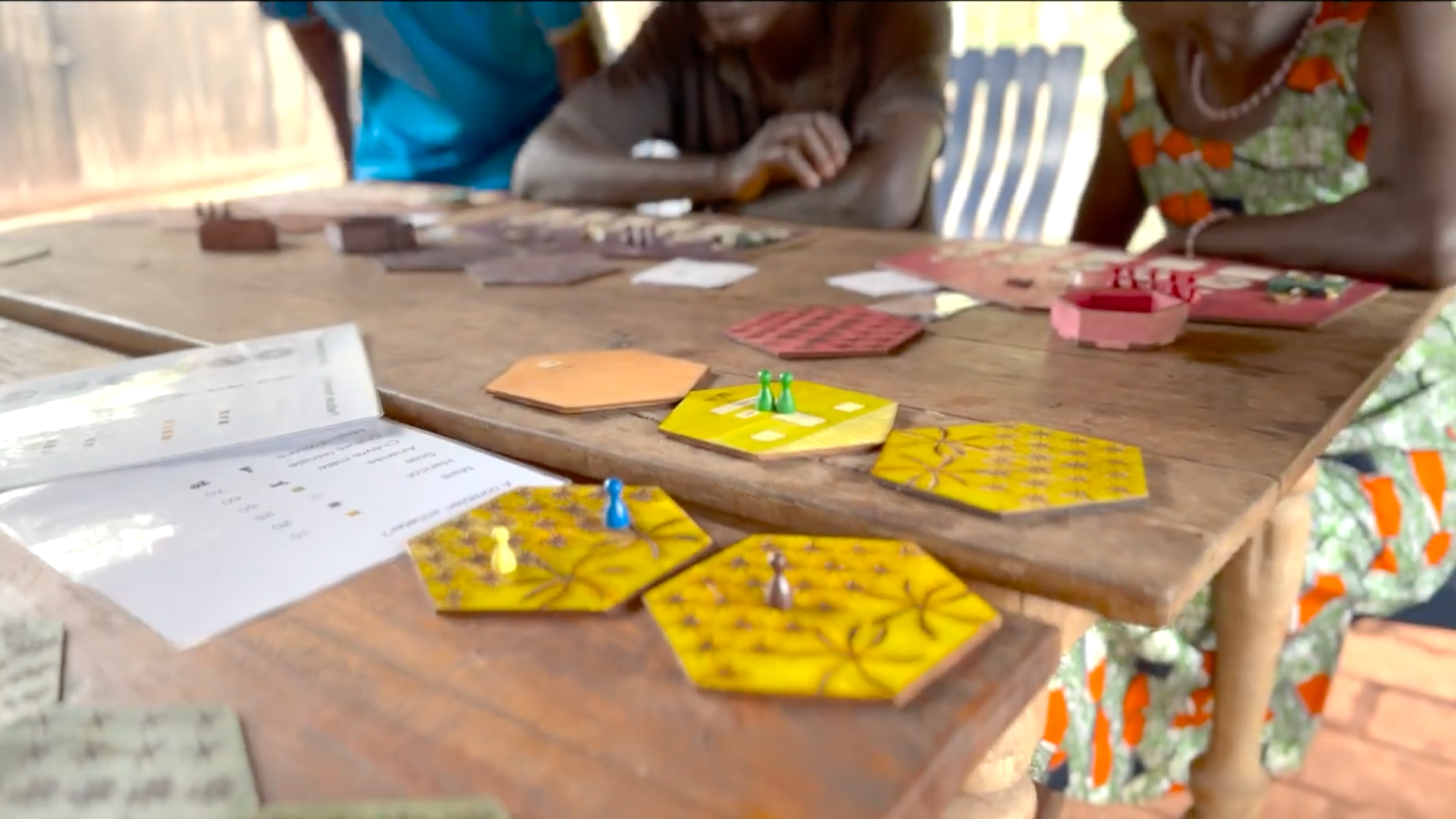 Yellow game pieces on a wooden table. In the background are farmers sitting at the table and studying the game. 